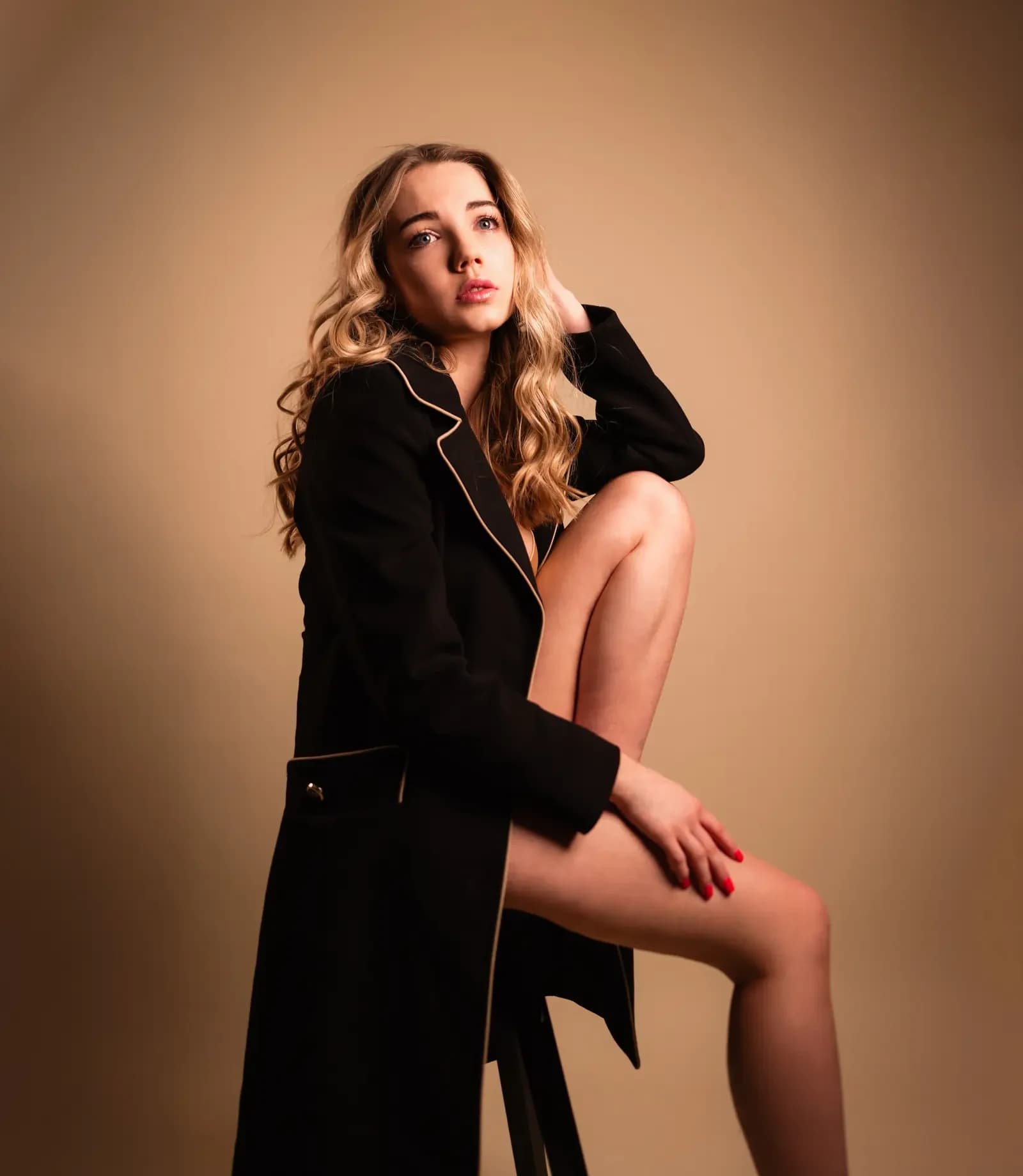 Model in schwarzem Mantel auf einem Hocker vor warmem, beigem Studiohintergrund